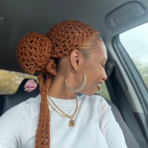 May include: A brown crochet head wrap with a large bun and long, braided ties. The head wrap is worn with gold hoop earrings and a gold necklace with a pendant. The person is wearing a white long-sleeved shirt.