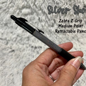 May include: A black Zebra Z-Grip retractable pen with a silver glitter barrel. The pen has a medium point and is labeled "Silver Shots".