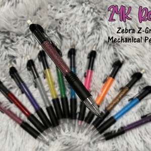 Könnte beinhalten: Eine Auswahl an Zebra Z-Grip Druckbleistiften mit glitzergefüllten Schäften in verschiedenen Farben, darunter Rot, Lila und Orange. Die Stifte haben schwarze Griffe und transparente Spitzen. Der Text auf dem Bild lautet "24K Rose" und "Zebra Z-Grip Mechanical Pencils."