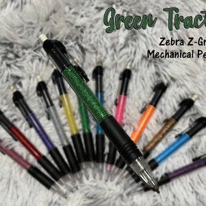 May include: A close-up of a green glitter Zebra Z-Grip mechanical pencil. The pencil is surrounded by other mechanical pencils in various colors, including pink, blue, purple, and yellow. The text "Green Tractor" and "Zebra Z-Grip Mechanical Pencils" is visible at the top of the image.