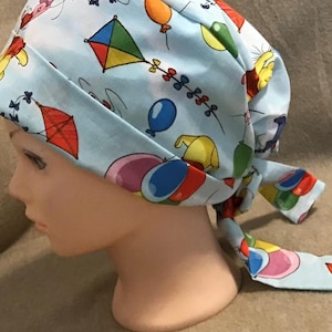 Könnte beinhalten: Hellblaue OP-Haube mit einem Muster aus Drachen, Luftballons und Zeichentrickfiguren. Die Haube wird hinten gebunden. Der Stoff hat ein verspieltes Design. Die Haube ist so konzipiert, dass sie den Kopf bedeckt.