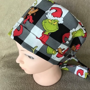 May include: A black and white checkered scrub hat with a repeating pattern of the Grinch wearing a Santa hat. The Grinch is green with a red and white Santa hat and a mischievous grin.