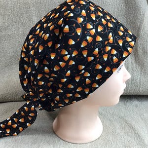 Puede incluir: Un gorro de quirófano negro con un patrón de maíz dulce naranja y blanco sobre un fondo negro. El gorro tiene una larga corbata que está atada en la parte posterior de la cabeza.
