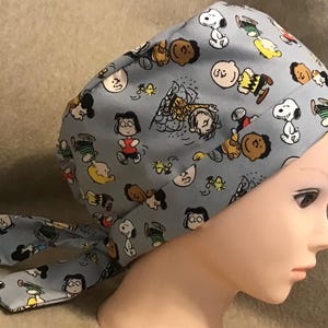 May include: A light blue surgical scrub cap featuring a repeating pattern of cartoon characters. The characters include Snoopy, Charlie Brown, and other figures from the Peanuts comic strip. The cap has a tie closure at the back.