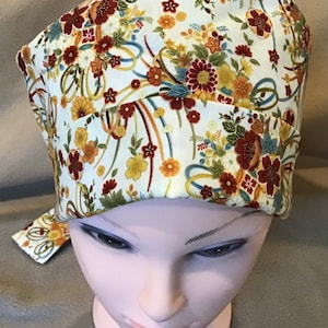 May include: A white scrub hat with a floral print featuring red, yellow, and green flowers. The hat has a tie on the back.