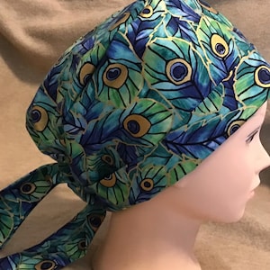 May include: A patterned scrub cap featuring a peacock feather design in shades of blue, green, and gold. The cap has a tie closure at the back and is made of a soft fabric. The cap is designed for medical professionals.