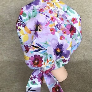 May include: A white scrub hat with a floral print featuring purple, pink, yellow, and green flowers. The hat has a long tie that can be tied at the back of the neck.
