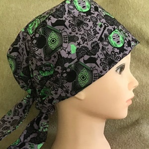 Könnte beinhalten: Eine violette und schwarze gemusterte OP-Mütze mit einem grünen und schwarzen Totenkopf- und Augenmotiv. Die Mütze hat zwei lange Bänder, die am Hinterkopf gebunden sind. Die Mütze hat den Text "1313" aufgedruckt.