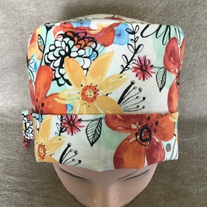 May include: A floral print hat with large orange, yellow, and pink flowers outlined in black on a white background. The hat has a folded brim and is displayed on a mannequin head. The design includes blue and green accents.
