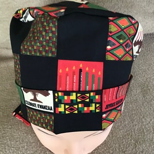 May include: A black fabric Kwanzaa hat with a colorful pattern of red, green, yellow, and black. The hat features the seven principles of Kwanzaa: Umoja (Unity), Kujichagulia (Self-determination), Ujima (Collective responsibility), Ujamaa (Cooperative economics), Nia (Purpose), Kuumba (Creativity), and Imani (Faith). The hat also includes the text "Celebrate Kwanzaa" and "Joyous Season".
