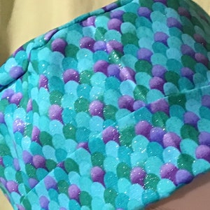 May include: A blue fabric scrub hat with a mermaid scale pattern in shades of green and purple. The fabric has a glittery finish.