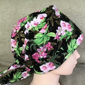 May include: A black fabric head covering with a pink and green floral pattern. The fabric has a gold metallic sheen. The head covering is tied at the back with a long tie.
