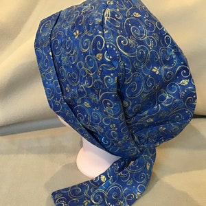 Könnte beinhalten: Eine blaue OP-Haube mit einem goldfarbenen Wirbelmuster. Die Haube wird am Rücken gebunden und besteht aus einem weichen Stoff. Das Design umfasst florale und Blatt-Akzente.