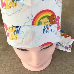 Könnte beinhalten: Weiße chirurgische Mütze mit einem Care Bears-Druck mit einem Regenbogen, Wolken und Sternen. Das Care Bears-Logo ist in der Mitte der Mütze sichtbar.