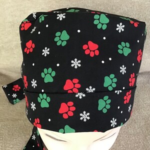 Op de afbeelding: Zwarte scrub-muts met een herhalend patroon van rode en groene poot afdrukken en witte sneeuwvlokken.