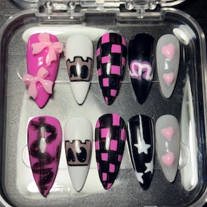 May include: A set of ten artificial nails in a clear plastic case. The nails feature various designs, including pink bows, a castle, a checkered pattern, a heart, and stars. The color palette includes pink, black, white, and gray.