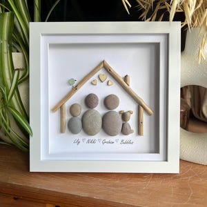 May include: A framed artwork featuring a family scene crafted from pebbles and twigs. The artwork depicts a house shape with a family of four and a small bird. The names Lily, Nikki, Graham, and Bubbles are written below.