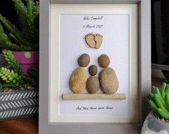 May include: Framed pebble art featuring a family of three, with a baby's footprints and the text "And then there were three." The artwork includes the name Niko Campbell and the date March 9, 2021. The frame is gray.
