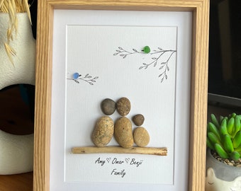 May include: Framed pebble art depicting a family of four, with two parents and two children, on a white background. The artwork includes a wooden frame, a branch, and the names "Amy", "Omar", and "Benji" with the word "Family" below.