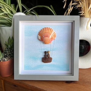 May include: A framed artwork featuring a seashell hot air balloon with two pebble people inside, on a blue and white watercolour background.