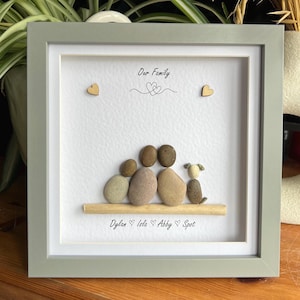 May include: A framed pebble art piece showing a family of five, with a dog, on a white background. The text "Our Family" is above the figures. The names Dylan, Isla, Abby, and Spot are below. The frame is a pale green.