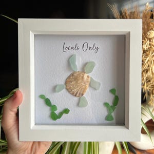 May include: White framed artwork with a sea shell and sea glass design. The artwork features a central shell with light green sea glass pieces. The text "Locals Only" is written at the top of the artwork.