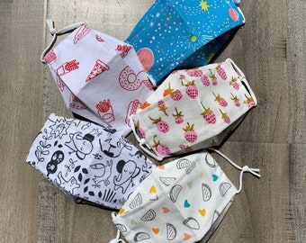 May include: Five different fabric face masks with various patterns. The masks are white with different colored designs. One mask has a blue background with white stars, another has a red and white design with a pizza slice, another has a red and white design with strawberries, another has a black and white design with a cartoon animal, and the last one has a white background with a red and white design of hearts and triangles.