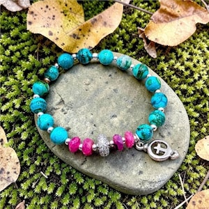 May include: A beaded bracelet featuring round turquoise-coloured stones with dark veining, interspersed with small silver-toned spacers. The design includes four vibrant pink beads, a central textured silver-toned bead, and a silver-toned cross charm.
