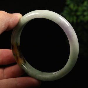 May include: A jade bangle bracelet with a smooth, polished surface. The bracelet features a blend of colors, including pale green, lavender, and hints of brown. The circular design is held by a hand, showcasing the bracelet's shape and color variations.