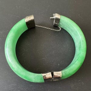May include: A green jade bangle bracelet with a silver clasp. The bracelet is made of a smooth, polished jade stone and has a simple, elegant design.