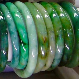 May include: A stack of jade green bangle bracelets with a glossy finish. The bracelets are stacked on top of each other, with the top bracelet being the most visible. The bracelets are all different sizes and have a slight variation in color.