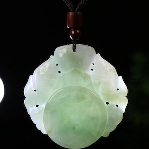 May include: A jade pendant with a carved floral design and a central circular element. The pendant is light green and white, with a dark brown cord and wooden bead for hanging. The background is black.