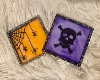 Puede incluir: Dos posavasos cuadrados de resina con bordes de purpurina. Un posavasos es naranja con una telaraña negra y arañas. El otro posavasos es morado con una calavera y huesos cruzados negros.