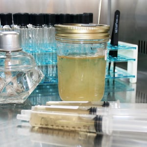 May include: A laboratory setting with a clear glass jar filled with a yellow liquid, a clear glass bottle with a silver lid, and three clear plastic syringes filled with a yellow liquid. The jar and bottle are on a stainless steel surface.