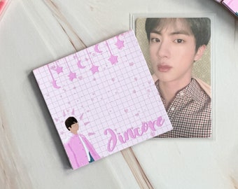 Può includere: Blocco note rosa e bianco con un motivo a griglia e un'illustrazione di cartone animato di una persona con una camicia rosa e la parola "Jincore" scritta in corsivo. Il blocco note ha un design di stelle e mezze lune.