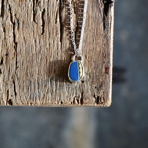 May include: A silver necklace with a pendant featuring a blue and cream-colored design. The pendant is a unique shape and hangs from a delicate chain. The necklace is displayed on a weathered wooden surface.