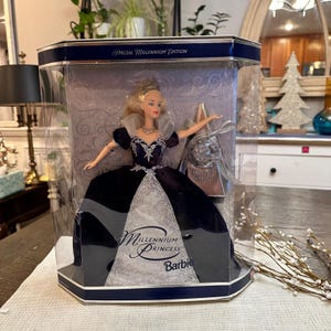 May include: A Millennium Princess Barbie doll in a clear display case. The doll wears a dark blue gown with a silver bodice and accents. The packaging reads "Special Millennium Edition" and "Millennium Princess Barbie".