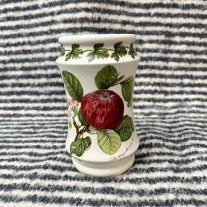 May include: A white ceramic vase with a botanical design featuring a large red apple, green leaves, and white blossoms. The vase has a flared shape and a decorative border of green leaves. The text "The Berry Morning" is printed on the vase.