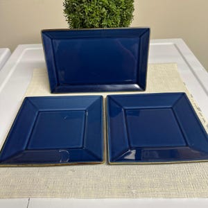 May include: A set of three navy blue ceramic plates. Two are square, and one is rectangular. Each plate has a raised border and a contrasting tan rim. The plates are arranged on a neutral-colored placemat.
