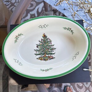 May include: An oval ceramic serving bowl with a cream-colored base and a green rim. The center features a detailed illustration of a decorated Christmas tree with gifts underneath, surrounded by small sprigs of holly and berries along the edge.