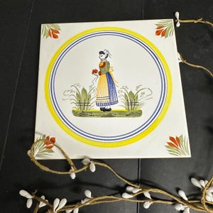May include: Square ceramic tile with a hand-painted illustration of a woman in traditional attire holding flowers. The design is framed by a yellow and blue circular border. Red and green floral accents are in each corner.