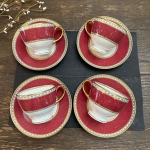 May include: A set of four porcelain teacups and matching saucers featuring a deep red textured pattern, white lower sections, and gold trim. Each cup has a curved gold handle and a pedestal base, while the saucers display a matching red border.