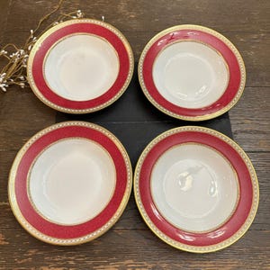 May include: A set of four round ceramic bowls featuring a wide, textured crimson red rim with a fine gold-tone border and a central white basin. The bowls are arranged in a two-by-two grid, showcasing a consistent design with a smooth finish.