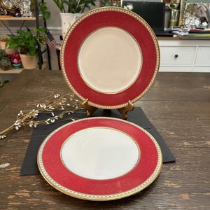 May include: Two round ceramic plates featuring a solid cream-colored center and a wide, textured red border. The rim is decorated with a delicate white and gold geometric pattern, providing a refined contrast to the vibrant red edge of the plates.