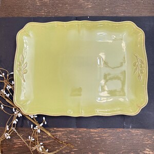 May include: A rectangular, light green ceramic serving tray with a scalloped edge and a glossy finish. The tray features embossed olive branch motifs on the left and right sides, adding subtle texture to the smooth, flat surface of the dish.