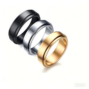 Può includere: Tre anelli in metallo nero, argento e oro. Ogni anello ha una fascia centrale che ruota liberamente all'interno dell'anello esterno. Gli anelli sono esposti su uno sfondo bianco, mettendo in risalto le loro superfici lucide.