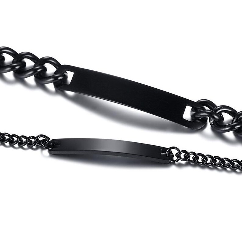 May include: Two black metal bracelets featuring smooth, rectangular identification plates. One bracelet consists of thick, interlocking curb chain links, while the other is composed of a finer, delicate cable chain. Both pieces have a matte finish.