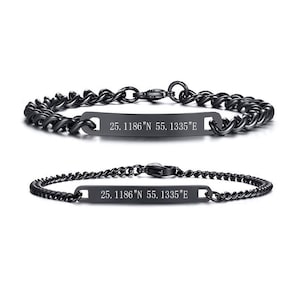 May include: Two black metal chain bracelets featuring a central rectangular bar. Each bar is engraved with the coordinates 25. 1186"N 55. 1335"E. The bracelets have a lobster claw clasp and a polished dark finish, showcasing a link chain design.