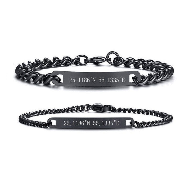May include: Two black metal chain bracelets featuring a central rectangular bar. Each bar is engraved with the coordinates 25. 1186"N 55. 1335"E. The bracelets have a lobster claw clasp and a polished dark finish, showcasing a link chain design.