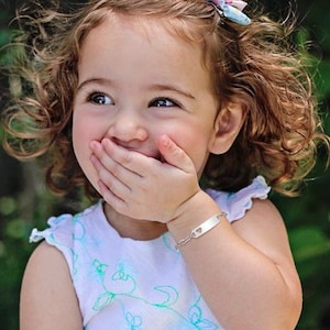 May include: A young girl with curly brown hair is smiling and covering her mouth with her hand. She is wearing a white shirt with a floral pattern and a silver bracelet with a heart-shaped charm.